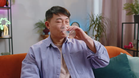 Thirsty-asian-man-sitting-at-home-holding-glass-of-natural-aqua-make-sips-drinking-still-water