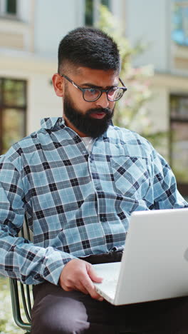 Indian-happy-man-working-on-laptop-celebrate-success-win-money-sitting-on-urban-street-in-city