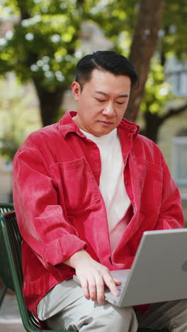 Asian-man-freelancer-sitting-on-city-street-opens-laptop-start-working-online-distant-job-outdoors