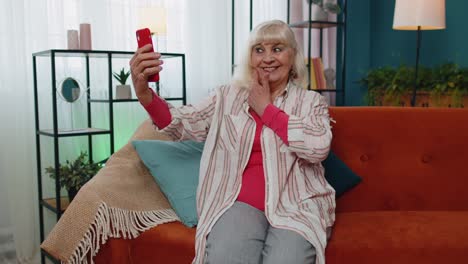 Happy-senior-grandmother-holding-smartphone-watching-mobile-video-calling-online-making-selfie-home