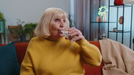 Thirsty-grandmother-woman-sits-at-home-holding-glass-of-natural-aqua-make-sips-drinking-still-water