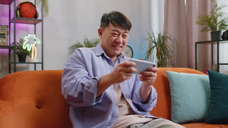 Worried-asian-handsome-man-playing-racing-or-shooter-video-games-on-smartphone-at-home-on-couch