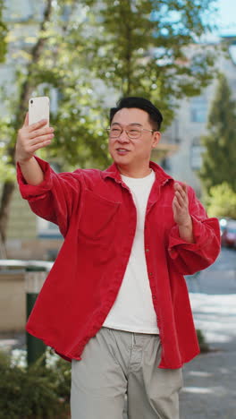 Asian-man-blogger-taking-selfie-on-smartphone-video-call-online-with-subscribers-on-city-street