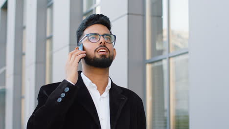 Indian-businessman-having-remote-conversation-talking-on-smartphone,-good-news-gossip-in-city-street