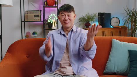 Asian-man-smiling-friendly-at-camera,-waving-hands-gesturing-hello,-hi,-greeting-at-home-on-sofa