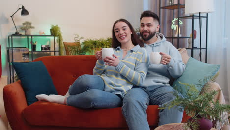 Smiling-family-couple-man-woman-drinking-a-cup-of-coffee-or-herbal-tea-at-home-sofa-in-the-morning