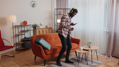 African-American-young-man-in-headphones-listening-music-dancing,-singing-in-living-room-at-home