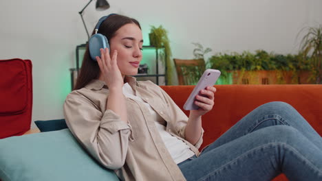 Happy-young-woman-in-wireless-headphones-relaxing-lying-on-sofa-at-home-listening-favorite-music