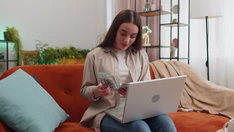 Una-Mujer-Feliz-Y-Sonriente-Contando-Dinero-En-Efectivo-Y-Usando-Una-Computadora-Portátil-Para-Calcular-Los-Ingresos-Domésticos-En-Casa