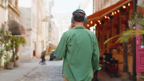 Rear-view-of-happy-relaxed-man-tourist-in-wireless-headphones-listening-music-walking-on-city-street