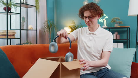 Unhappy-man-unpacking-carton-box-feeling-dissatisfied-with-getting-damaged-wireless-headphones-home