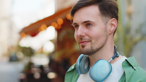 Thoughtful-happy-smiling-male-tourist-with-wireless-headphones-around-neck-looking-up-on-city-street