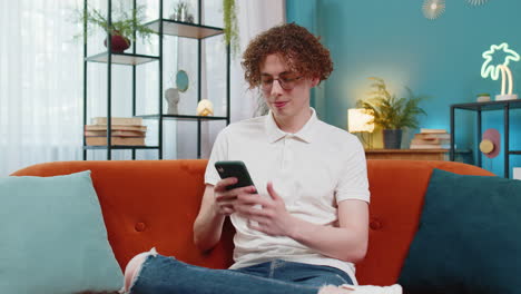 Caucasian-young-man-sitting-on-couch-using-mobile-smartphone-share-messages-smiles-at-home-room