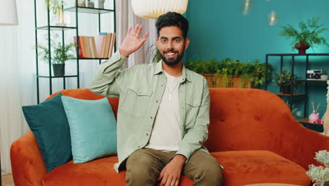 Indian-man-smiling-friendly-at-camera,-waving-hands-gesturing-hello,-hi,-greeting-at-home-on-sofa