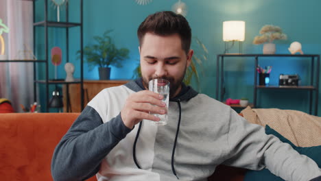 Thirsty-Caucasian-man-sitting-at-home-holding-glass-of-natural-aqua-make-sips-drinking-still-water