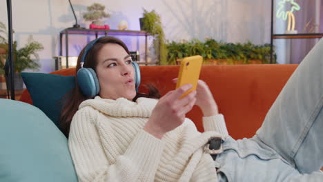 Happy-young-woman-in-wireless-headphones-relaxing-lying-on-sofa-at-home-listening-favorite-music