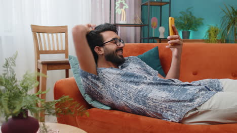 Indian-hindu-young-man-lying-on-couch,-using-smartphone-share-messages-on-social-media-application