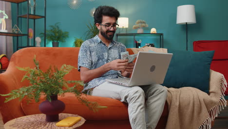 Un-Hombre-Indio-Sonriente-Contando-Dinero-En-Efectivo-Y-Usando-Una-Computadora-Portátil-Para-Calcular-Los-Ingresos-Internos-En-Casa