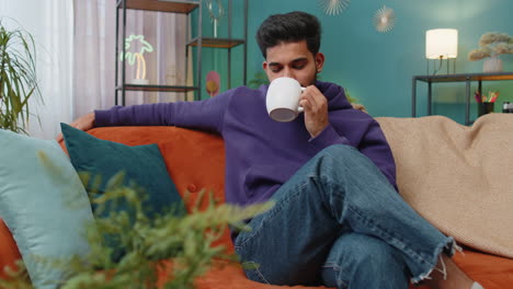 Handsome-smiling-indian-man-drinking-a-cup-of-coffee-or-herbal-tea-sitting-at-home-couch,-break-time