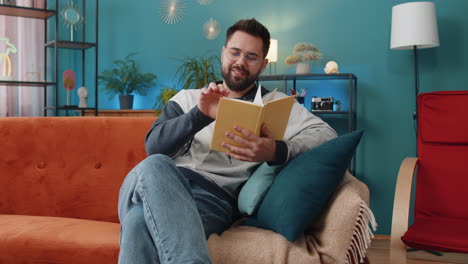 Young-man-relaxing-interesting-book-turning-pages-smiling-enjoying-literature-taking-a-rest-on-sofa