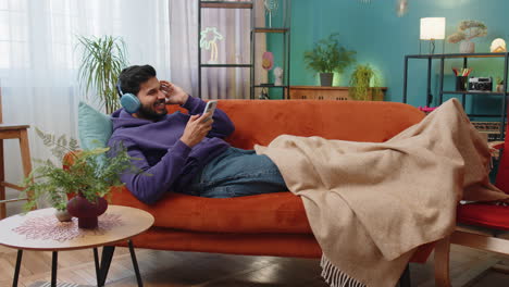 Happy-indian-man-in-wireless-headphones-relaxing-sitting-on-sofa-at-home-listening-favorite-music