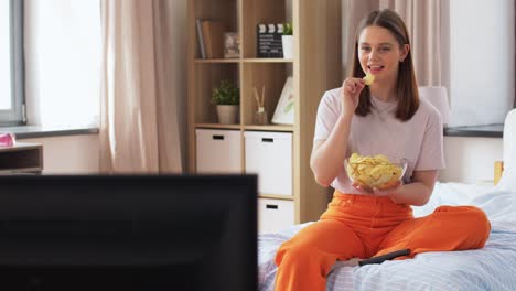 Glückliches-Mädchen-Mit-Chips-Vor-Dem-Fernseher-Zu-Hause.-Ungesunde-Ernährung,-Fast-Food-Und-Menschen-Konzept-Glückliches-Mädchen-Mit-Fernbedienung-Und-Chips-Vor-Dem-Fernseher-Sitzend-Auf-Dem-Bett-Zu-Hause