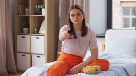 Happy-Girl-with-Crisps-Watching-Tv-at-Home.unhealthy-eating,-fast-food-and-people-concept-happy-girl-with-remote-control-and-crisps-watching-tv-sitting-on-bed-at-home