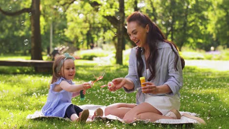 Mother-with-Daughter-Blowing-Soap-Bubbles-at-Park.family,-motherhood-and-people-concept-happy-mother-with-little-daughter-blowing-soap-bubbles-at-summer-park-or-garden