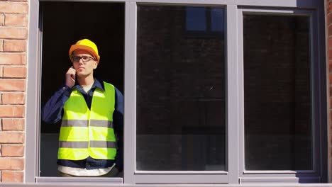 Builder-Looking-Out-Window-and-Calling-on-Phone.construction-business-and-building-concept-male-builder-in-helmet-and-safety-west-calling-on-smartphone-and-looking-out-window