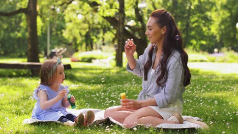 Mother-with-Daughter-Blowing-Soap-Bubbles-at-Park.family,-motherhood-and-people-concept-happy-mother-with-little-daughter-blowing-soap-bubbles-at-summer-park-or-garden