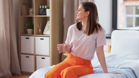 Girl-with-Glass-of-Water-Sitting-on-Bed-at-Home.people-concept-happy-smiling-girl-with-glass-of-water-sitting-on-bed-at-home