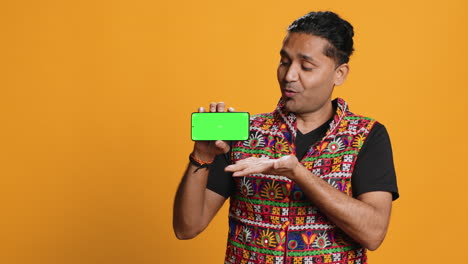 Smiling-man-presenting-cellphone-with-green-screen-display,-studio-background