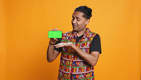 Smiling-man-presenting-cellphone-with-green-screen-display,-studio-background