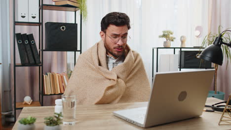 Sick-ill-Indian-businessman-wrapped-in-blanket-works-on-laptop-at-home-office-desk-takes-medicine