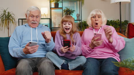 Granddaughter-and-grandparents-celebrating-victory-win-online-game-lottery-on-smartphones-at-home