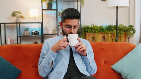 Tired-smiling-Indian-man-drinking-cup-of-warm-coffee-or-herbal-tea-in-the-morning-relaxing-enjoying