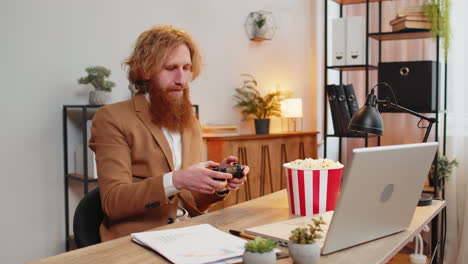 Businessman-eating-popcorn-while-playing-video-game-on-laptop,-spending-leisure-time-at-home-office