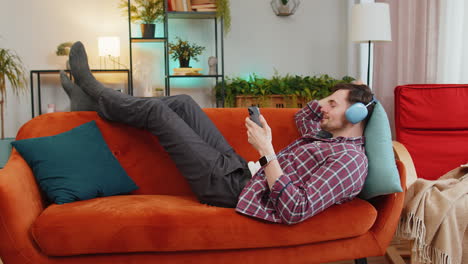 Man-in-wireless-headphones-listening-energetic-dancing-music-on-smartphone-relaxing-lying-on-sofa