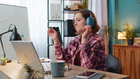 Excited-business-woman-listening-music-through-wireless-headphones-sitting-at-home-office-desk