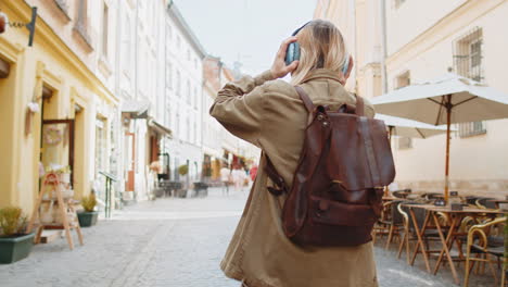 Rear-view-of-young-woman-tourist-with-backpack-listening-music-via-headphones-walking-through-street