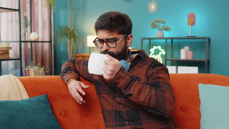 Attractive-smiling-Indian-young-man-drinking-a-cup-of-coffee-or-herbal-tea-at-home-couch-in-morning