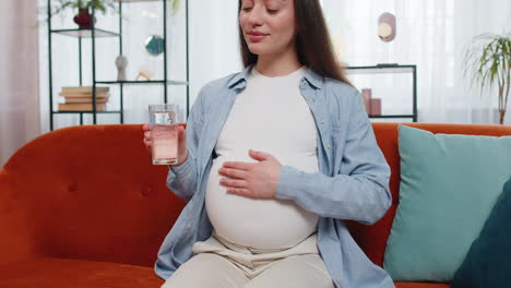 Pregnant-woman-holding-glass-with-water-and-drinking-while-sitting-on-sofa-healthy-maternity-concept