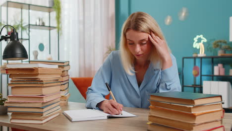 Tired-young-student-writer-woman-sitting-at-desk-with-pile-of-books-writing-a-novel-book-with-pen