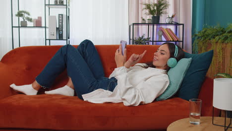 Mujer-Joven-Feliz-Con-Auriculares-Inalámbricos-Relajándose-Tumbada-En-El-Sofá-De-Casa-Escuchando-Su-Música-Favorita