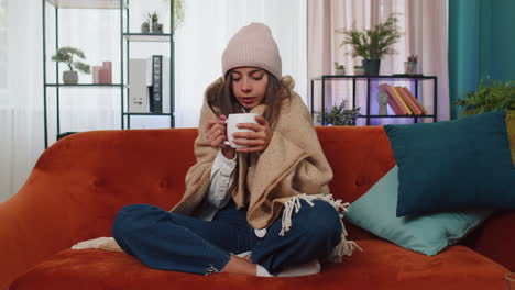 Sick-young-woman-wear-hat-wrapped-in-plaid-sit-alone-shivering-from-cold-on-sofa-drinking-hot-tea