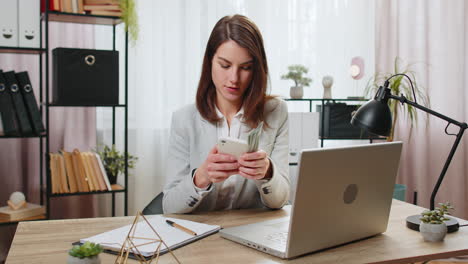 Happy-rich-business-woman-counting-money-cash-dividends-using-smartphone-calculator-in-home-office