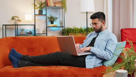 Indian-man-typing-working-on-laptop-using-apps-browsing-internet-lying-study-education-on-home-sofa