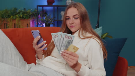 Smiling-happy-young-woman-at-home-counting-money-cash-use-smartphone,-income,-saves,-lottery-win