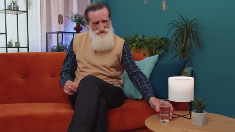 Thirsty-senior-old-man-sitting-at-home-holding-glass-of-natural-aqua-make-sips-drinking-still-water