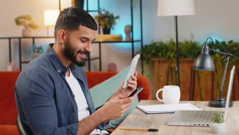 Rich-Indian-man-freelancer-using-smartphone-counting-money-cash-calculate-earnings-at-home-office